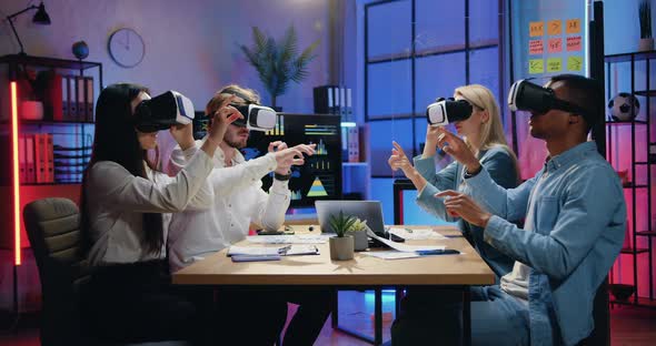Team of Coworkers Sitting Around the Table in Evening Office and Working in Virtual Reality Headsets