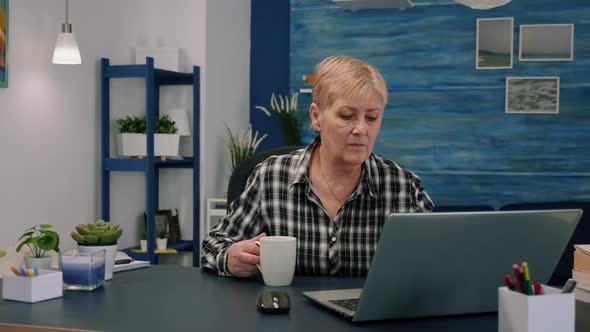 Old Businesswoman Sitting at Workplace Typing at Laptop Drinking Coffee