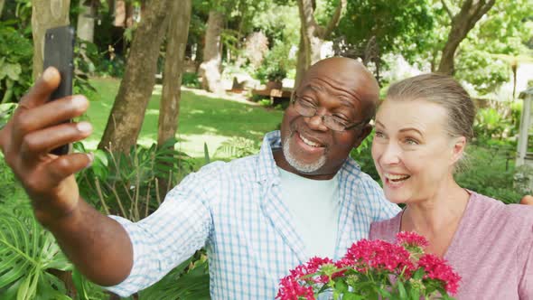 Happy senior diverse couple wearing shirts and taking selfie with smartphone in garden