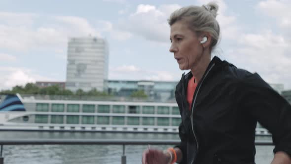 Mature Woman in Earphones Running on Street Near City River