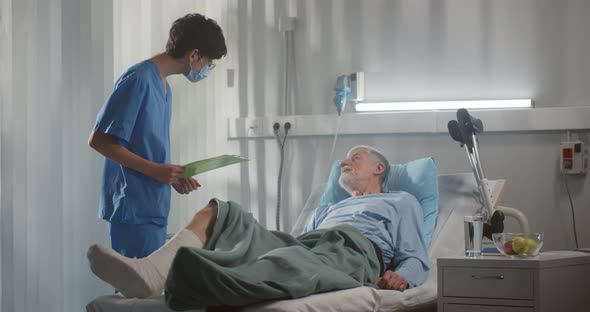 Surgeon in Medical Mask Examining Aged Male Patient with Broken Leg