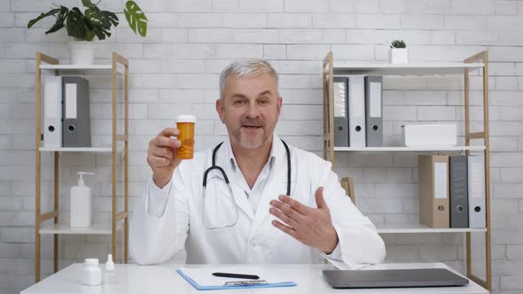 Mature Doctor Showing Tube With Medical Pills Sitting In Office