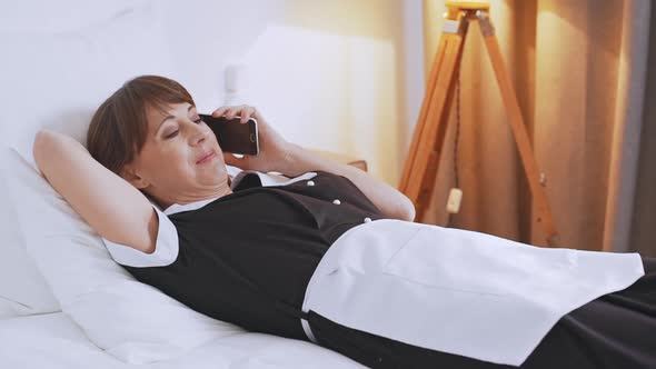 Female Middleaged Caucasian Chambermaid Lying on Bed in Umiform Talking on Mobile Phone in