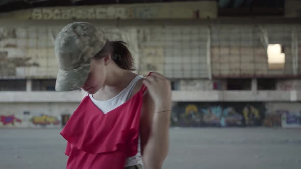 Cute Young Woman in Military Uniform Trying on Red Dress and Dancing Around, Smiling in Abandoned