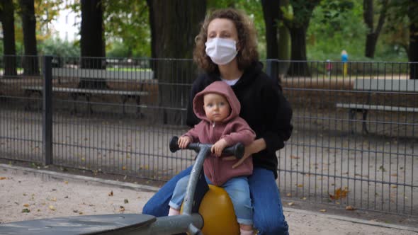 Woman in Protective Mask and Small Child Play Alone on Balancer in Green Park Area During Lockdown