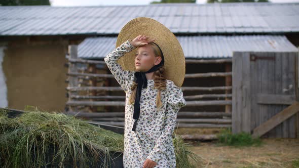 Beautiful Girl with Two Braids in Hat and Romantic Dress Looking Away on Rural Landscape in