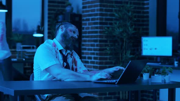 Scary Looking Zombie Trying to Work on Modern Laptop in Office Workspace at Night