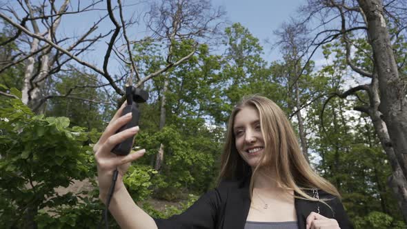 Young Woman With Small Gimbal With Camera Making Selfie Video in Green Forest on Sunny Summer Day, T