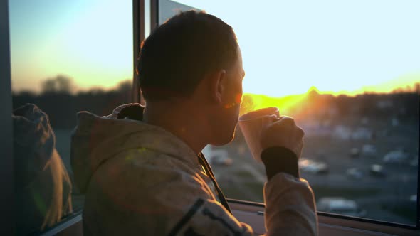 A young man in a sweatshirt drinks hot tea by the open window during a beautiful sunset