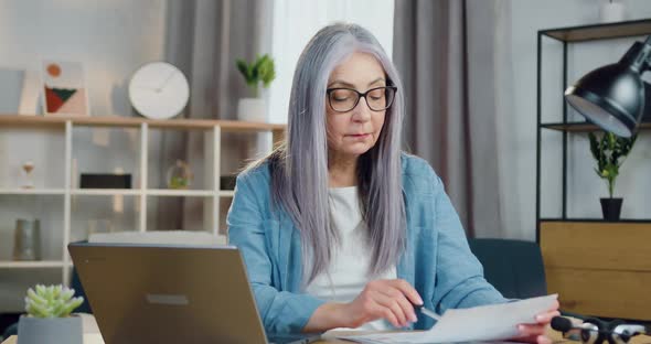 Woman Sitting in front of Computer and Checking Paper Report with Datas on Laptop Screen