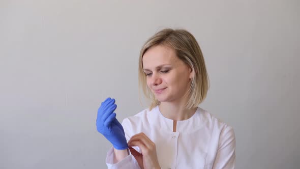 Blonde Female Doctor Surgeon in White Uniform Puts on Blue Gloves and Medical Mask