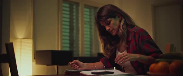 Young woman sitting at desk, studying at home in the evening, making notes 