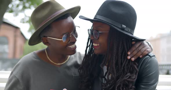 Happy Young Affectionate Black Couple in Stylish Wear Sitting on Bench Using Smartphone