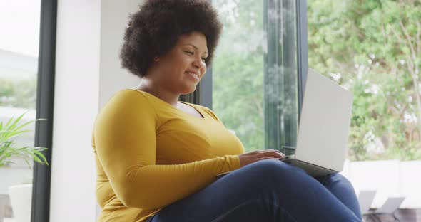 Video of happy plus size african american woman sitting on floor and using laptop