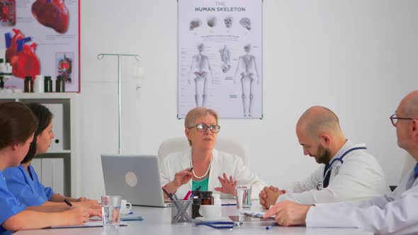 Elderly Specialist Woman Doctor Sitting on Conference Desk Talking with Team