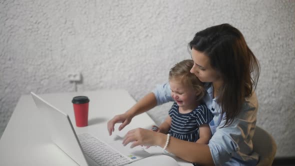 Woman Distracting a Child and Continues Work on Notebook. Young Woman Calms Crying Child in Office.