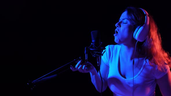 Caucasian Woman in Headphones Singing Into a Microphone in Neon Light on a Black Background