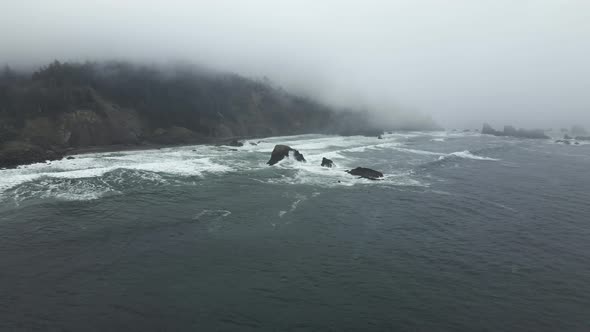 Fog shrouds steep cliffs as white foam and emerald waves crash along a jagged coastline, aerial orbi