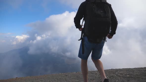 Young Male Hiker with Backpack Reaching Up Top of Mountain with Cloudy Sky at Background and Raising