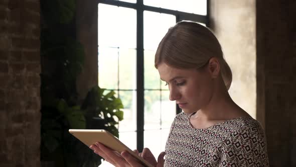 Young attractive smiling caucasian woman uses tablet with window background.