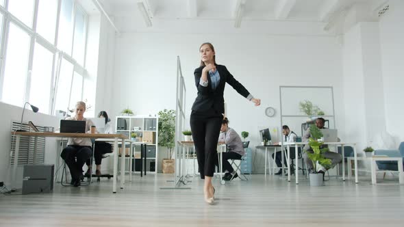 Elegant Young Woman Dancing in Workplace Moving to Music While Employees Working at Desks