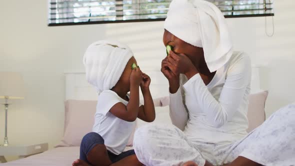 Happy african american mother and daughter wearing towels sitting on bed putting cucumbers on eyes