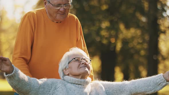 Elderly Couple in the Park. Husband Pushing His Disabled Wife with Outstretched Hands in the