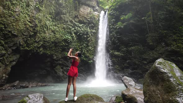 A Young Tourist Standing at a Waterfall Capturing Photos Using a Smartphone