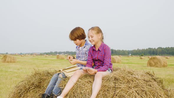 Teenager Boy and Girl Sitting on Haystack at Countryside Field. Carefree Boy and Girl Reading Book