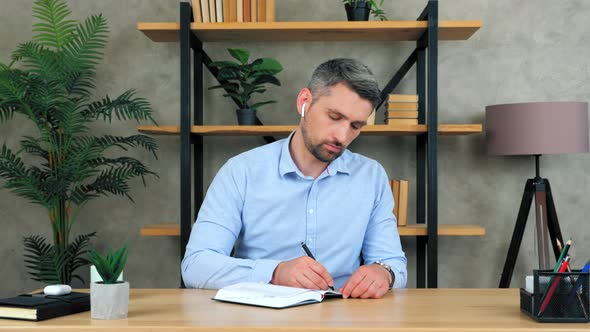 Serious confident businessman CEO in home office listening music writes information to notepad