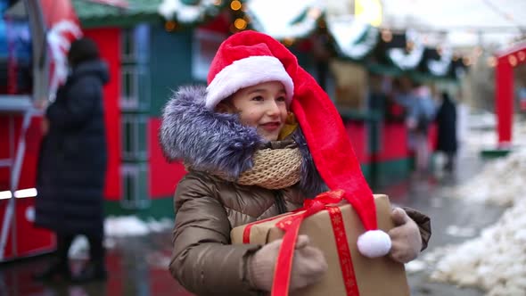 Portrait of joyful girl in Santa hat with gift box for Christmas on city street in winter with snow