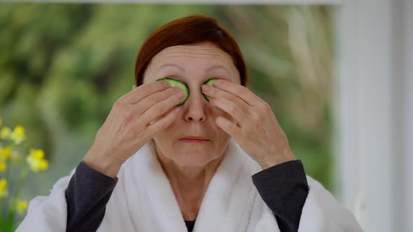 Front View Portrait of Mature Caucasian Woman Applying Cucumber on Eyes Standing at Home Indoors