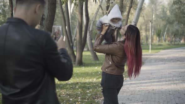 Young Caucasian Man Taking Photos of Smiling Beautiful Woman Playing with Infant in Sunny Park