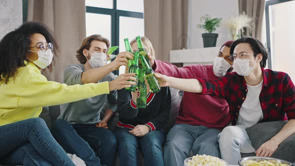 Happy Interracial Group of Friends in Protective Masks Clink Glasses with Beer Bottles While Sitting