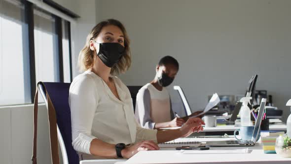 Two diverse female colleagues wearing face mask, working at desk in office