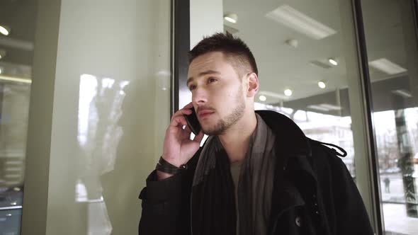 Concentrated Caucasian Young Handsome Male Standing Near Shop Window and Talking on Mobile Phone in