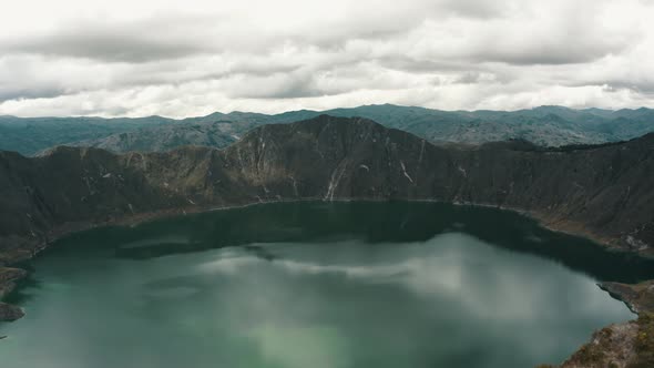Aerial backwards shot of natural crater lake surrounded by volcano rocks during cloudy day