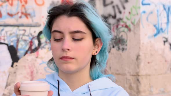 Blue haired teenage girl in oversize hoodie drinking coffee to go against graffiti wall