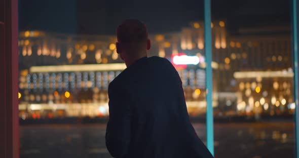 Back View of Businessman Celebrating Success Standing Near Panoramic Window with Night Cityscape