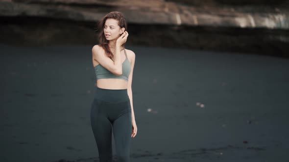 Sensual Woman in Sportswear Rests on Black Sand Beach Looks at Camera Smiles