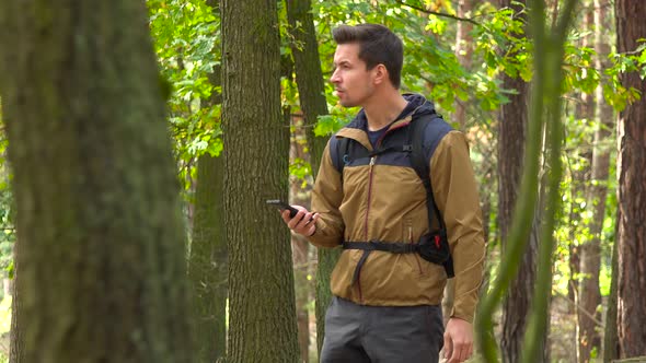 A Hiker Walks Through a Forest, Stops, Looks Around and on a Map on a Smartphone