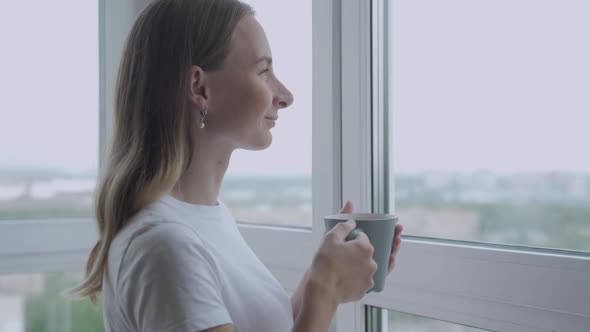 Woman Looking Out Window Enjoying Fresh New Day Feeling Rested and Drinking Coffee at Home. Slow