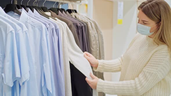 a Young Woman in a Protective Mask in a Clothing Store Chooses Casual Clothes Hung on Hangers