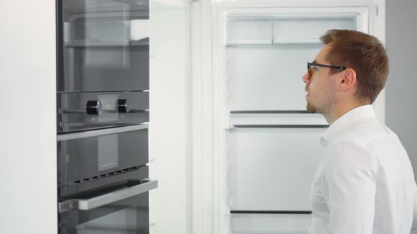 Side View of Young Guy in Glasses Opening Empty Fridge Looking for Dinner