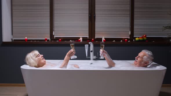 Senior Couple Grandfather and Grandmother Is Taking Foamy Bath, Drinking Champagne in Bathroom