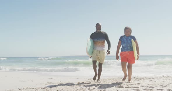 Happy senior african american couple walking with surfboards on sunny beach