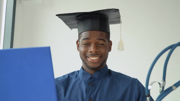 A Young African American Graduate Man in a Master's Hat and Blue Medical Gown Smiles Very Loudly