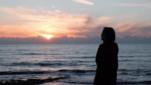 Young girl talking on phone at rocky shore at sunset with sun road reflection on sea water