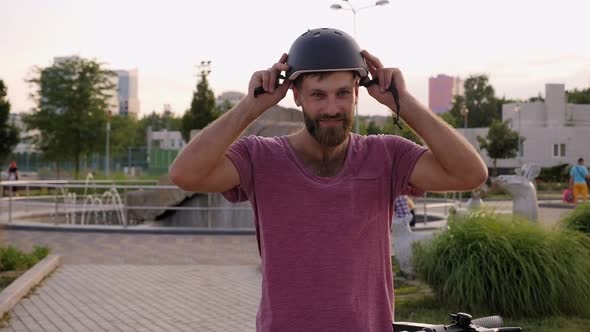 A Young Bearded Man Puts on a Protective Helmet in the Summer in the Park