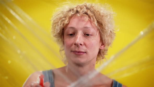 Closeup of Young Queer Man Spraying Water on Cling Film Standing at Yellow Background